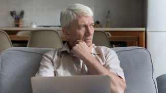Starší muž sedí na gauči, před sebou má notebook a tváří se zamyšleně