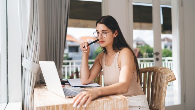 Žena sedící doma u notebooku s tužkou v ruce přemýšlí