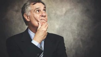 Starší muž v obleku stojí a drží se za bradu, kouká nad sebe a tváří se zamyšleně