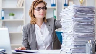 Mladá žena v brýlích sedí u stolu, vedle ní leží hromada dokumentu, které tápavě hledí
