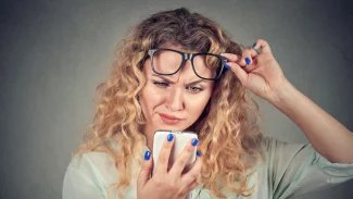 Mladá žena s brýlemi, které si drží na čele, kouká do mobilního telefonu a tváří se nechápavě
