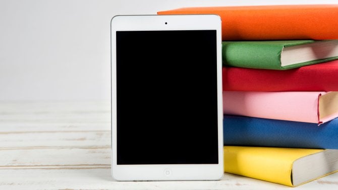 Moderní tablet spočívá vedle barevné hromádky knih na dřevěném povrchu pro studium a volnočasové aktivity