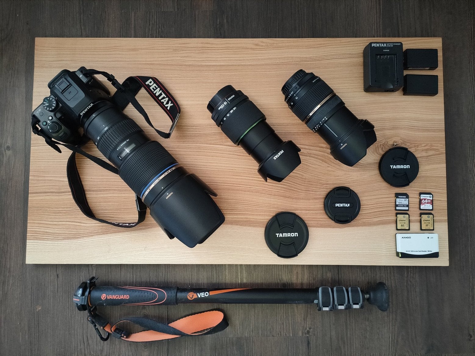 Digitální zrcadlovka Pentax K-S2 s nasazeným objektivem Tamron AF SP 70-200mm f/2,8 Di LD IF Macro s nasazenou sluneční clonou a dále objektiv  Pentax SMC DA 18 – 135 mm f/3.5 – 5.6 ED AL (If) DC WR a úplně vpravo je TAMRON 28-75 mm f/2,8 SP XR Di Asph. V pravém horním rohu fotografie je nabíječka s dvěma bateriemi a v pravém dolním rohu je čtečka paměťových karet a 4 paměťové karty standardu SDXC. Ve spodní části složený monopod.