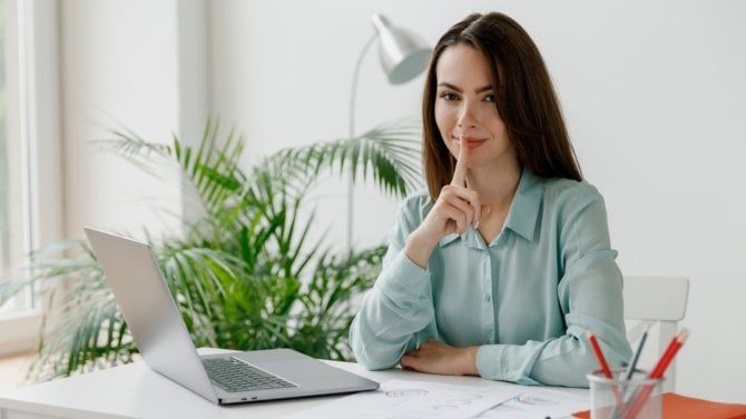 Žena sedí v kanceláři a má ruku před ústy jako znak tajemství