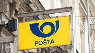 Na obrázku je tabule, na které je logo České pošty. Tabule je přidělaná na budově.