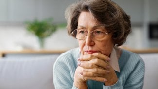 Žena v důchodovém věku v brýlích, sedí, má bradu v rukách a tváří se ustaraně