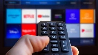 ruka drží ovladač k televizoru, v pozadí televize se streamovacími službami