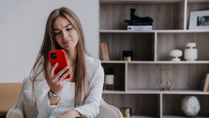 Frustrovaná mladá blondýna sedící na židli se dívá na telefon a s nešťastným výrazem ve tváři čte zprávu doma.