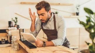 Mladý muž, číšník, stojí u pultu, v ruce drží ceduli a rozčiluje se nad ní