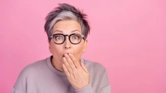 Šik starší žena s brýlemi a vykulenýma očima je v šoku s rukou před pusou