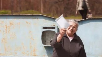 Seniorka stojí u poštovní schránky, v ruce drží dopis a prohlíží si jej proti sluníčku.