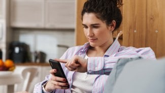 Tmavovlasá žena sedí s mobilním telefonem doma na gauči