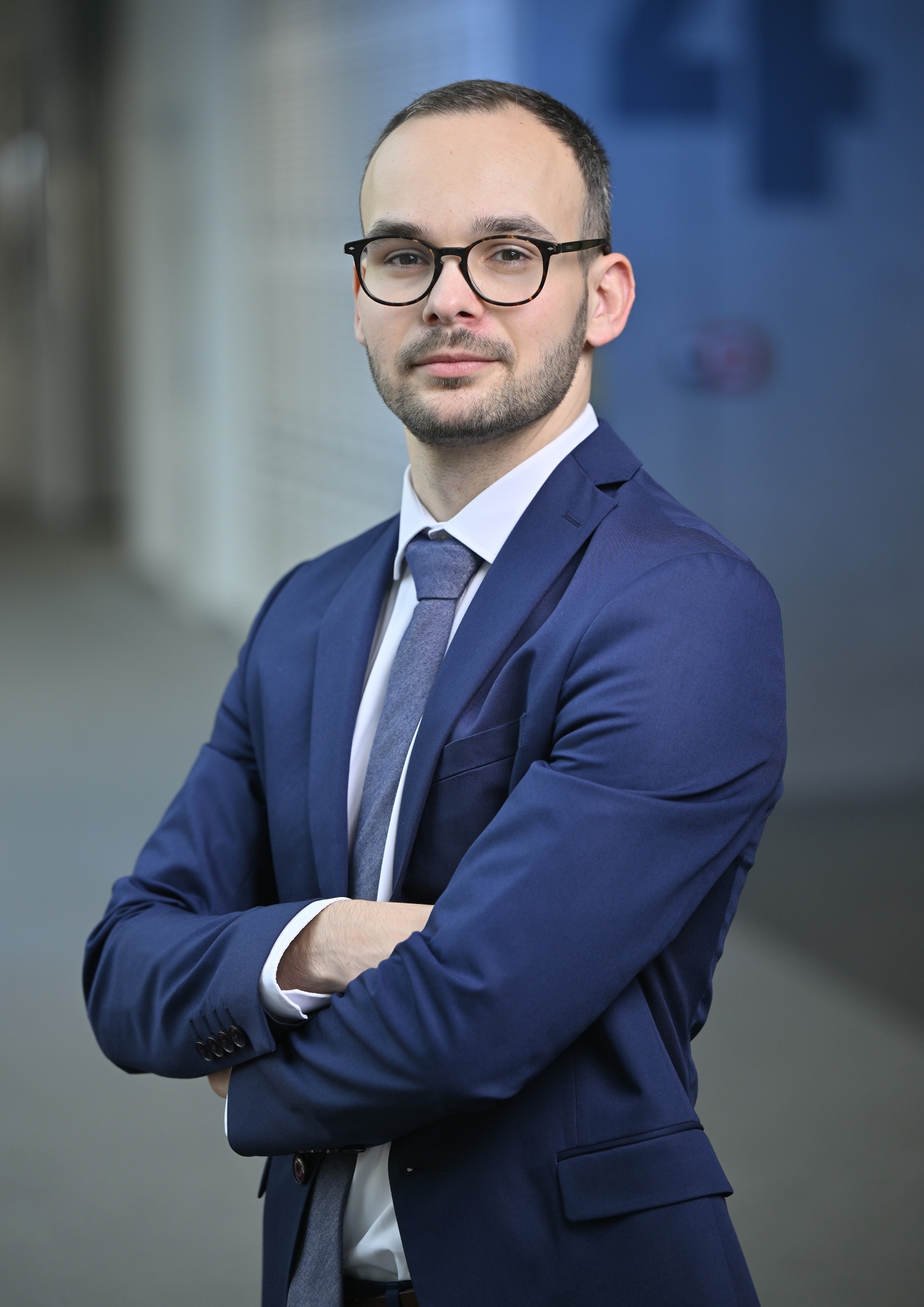 Muž v modrém obleku, kulatých brýlích, se zkříženýma rukama.