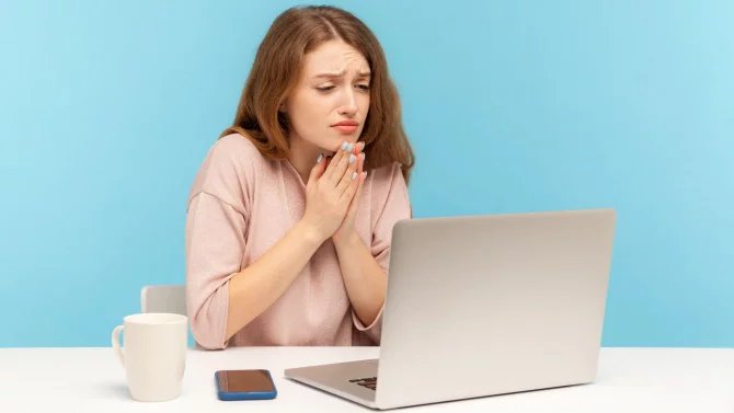 Žena sedící u notebooku má sepjaté ruce a smutný výraz