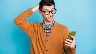 Mladý muž v brýlích drží v ruce mobilní telefon a tváří se překvapeně. Jednou rukou se drží za hlavu