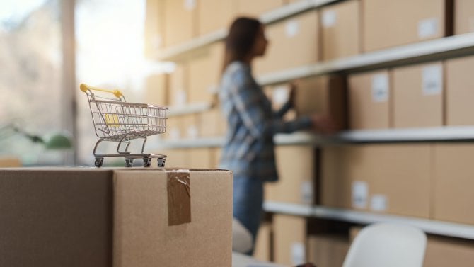 Majitel e-shopu přesouvá dodací krabice ve skladu, v popředí malý nákupní vozík: koncept online nakupování a doručování