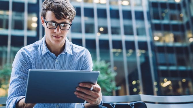 Úspěšný mužský obchodník v módním formálním oblečení čtení finančních zpráv na internetových stránkách pomocí 4G internetu na moderním tabletu sedí venku na ulici obchodního města s moderními mrakodrapy