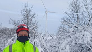 Mladý muž v pracovním oděvu, celý zahalený, stojí v prostoru před větrnou elektrárnou, která se tyčí v zamrzlé přírodě