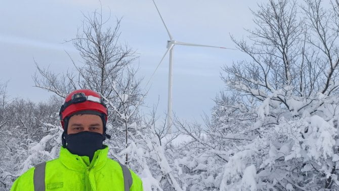 Mladý muž v pracovním oděvu, celý zahalený, stojí v prostoru před větrnou elektrárnou, která se tyčí v zamrzlé přírodě