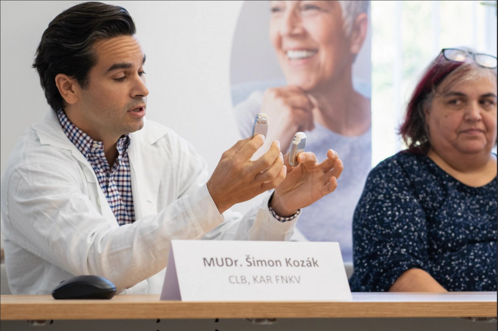Šimon Kozák ukazuje, o kolik je baterie nového stimulátoru menší (drží ji v pravé ruce) než baterie dosud používaných přístrojů. Prezentaci sledovala také pacientka Marika Szabová, které lékař stimulátor voperoval v červnu 2024 (tehdy šlo o původní, nikoliv nový typ přístroje).