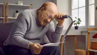 Starší muž sedí zoufale v křesle, opírá se o hůl, v druhé ruce drží dokument