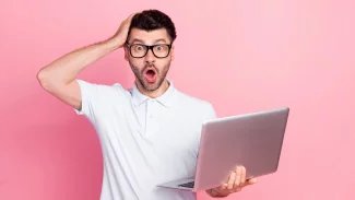 Mladý muž v brýlích stojí s počítačem v ruce a tváří se překvapeně