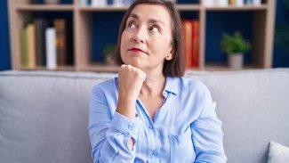 Starší žena sedí na gauči, kouká vzhůru a drží si pod bradou ruku v pstě