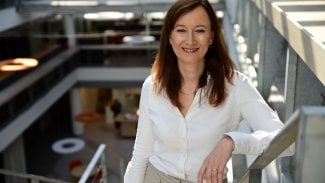 Žena v bílé košili stojí na schodech opřená o zábradlí.