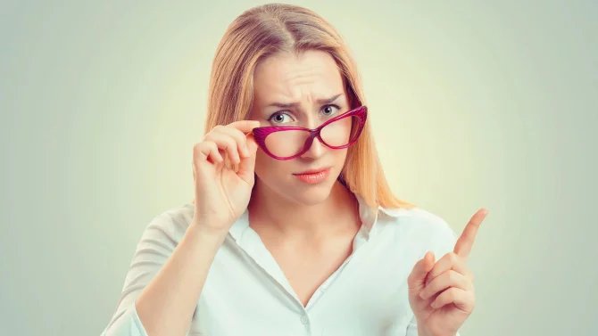 Mladá žena s brýlemi s upozorňujícím výrazem na nějaké povinnosti. Má zdvižený prst