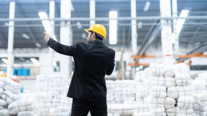 Muž v obleku s helmou, který stojí ve skladu a ukazuje rukou nahoru.