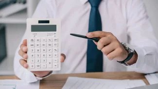 Muž v kravatě v kanceláři ukazuje perem na kalkulačku
