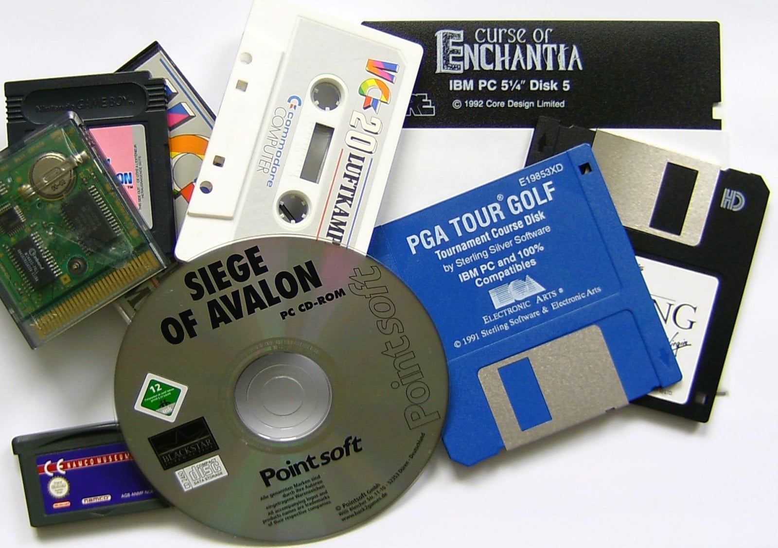 Kazeta, cartridge, diskety a herní CD-ROM rozložené na světlém podkladu.