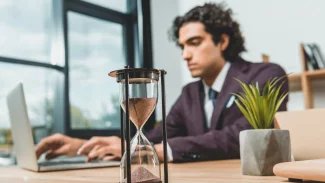 Mladý muž v pozadí obrázku sedí u počítače a pracuje. Před ním stojí přesýpací hodiny, které upozorňují na ubíhající čas.