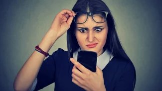 Mladá žena si drží brýle na čele a s údivem se dívá do mobilního telefonu