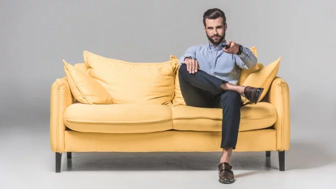 Mladý muž se dí na pohovce, drží v ruce ovladač a přepíná kanály na televizi