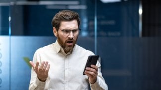 Mladý podnikatel ve stresu stojí v kanceláři a frustrovaně se dívá na svůj mobilní telefon a roztahuje ruce.