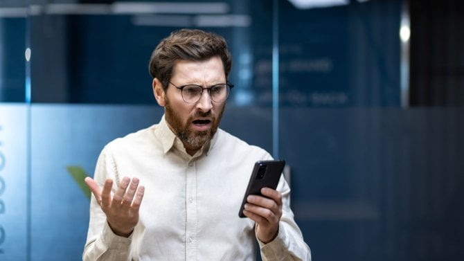 Mladý podnikatel ve stresu stojí v kanceláři a frustrovaně se dívá na svůj mobilní telefon a roztahuje ruce.