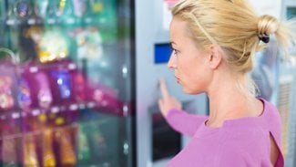 Mladá blonďatá žena stojí u automatu s drobnými sladkostmi a jinými potravinami a nakupuje.