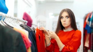 Mladá žena stojí u stojanu sv oblečením a prohlíží si cenovku. Tváří se rozčarovaně