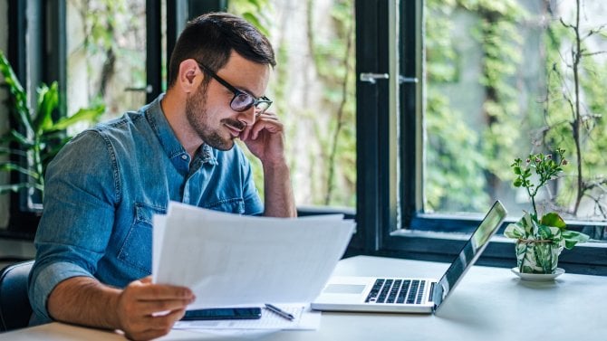 Mladý muž sedící u okna před notebookem na stole studuje papírové dokumenty
