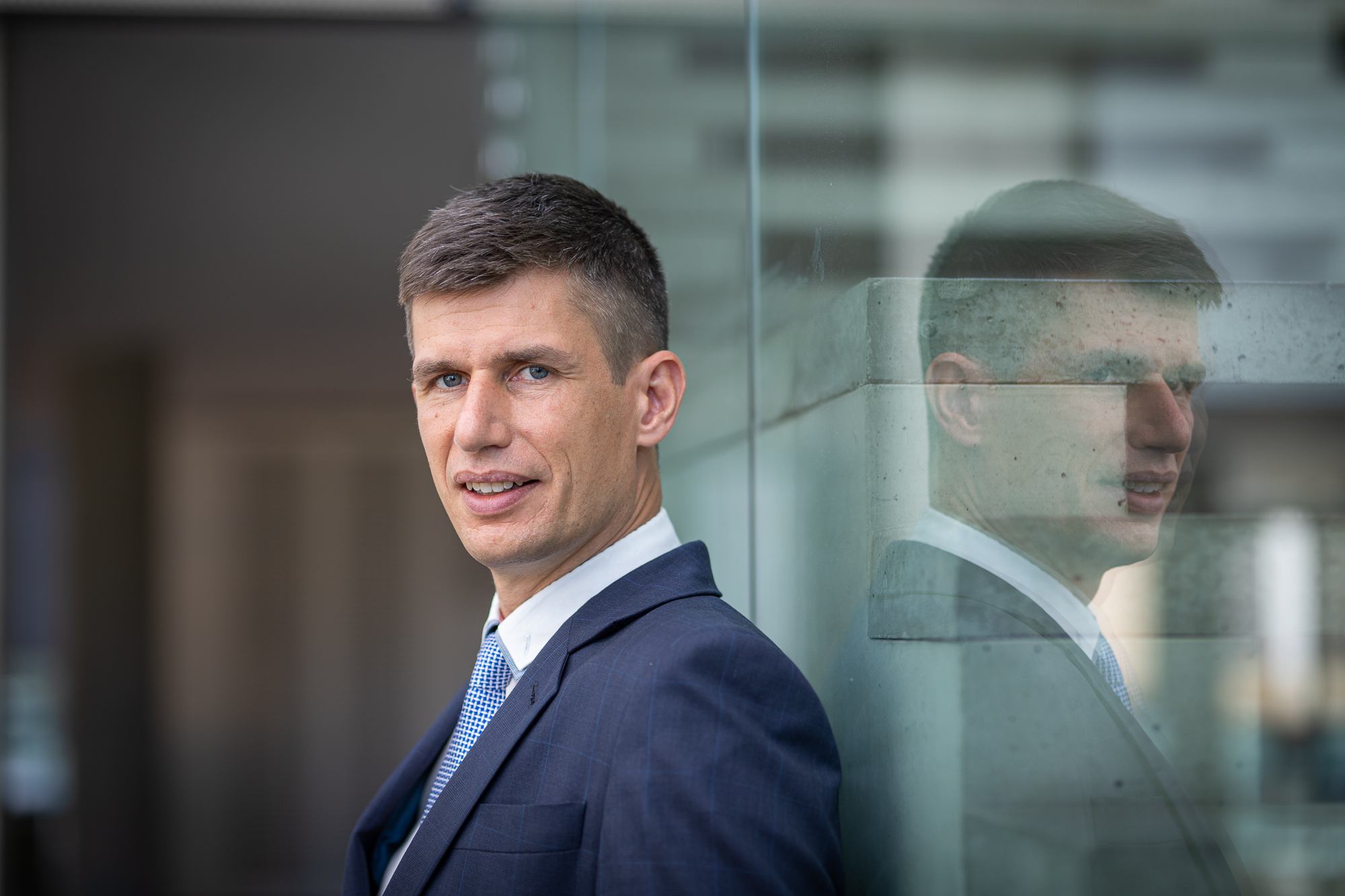 Fotografie muže v v modrém obleku, který stojí opřený o skleněnou stěnu, ve které se odráží jeho obličej.