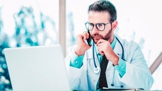 Mladý doktor sedí u stolu v bílém plášti, před sebou má notebooku a u ucha drží telefon