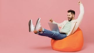 Mladý šťastný muž v módní bunda košile sedět v tašce židli držet používat práci na notebooku PC počítač dělat vítěze gesto izolované na obyčejné pastelové světle růžové pozadí studio.