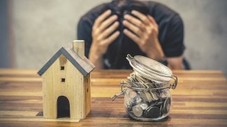 V popředí dřevěná maketa domu, sklenice s penězi a v pozadí muž držící se za hlavu.