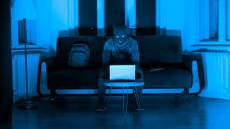 Zamyšleně vypadající zloděj se skrytou tváří, který používá notebook v tlumeně osvětlené místnosti, což naznačuje nelegální kybernetickou činnost.