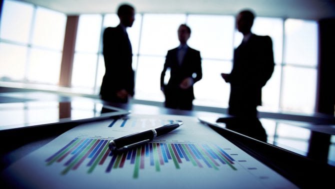 Lidé v kanceláři stojící vzadu ve stínu a v popředí je papír s perem