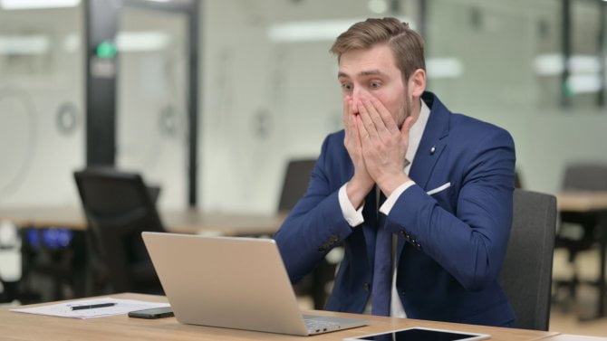 Překvapený muž sedící za stolem u notebooku si zakrývá ústa