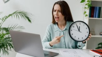 Žena u počítače s budíkem v ruce poukazuje na čas splnění termínu
