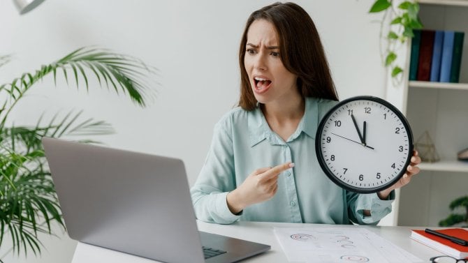 Žena u počítače s budíkem v ruce poukazuje na čas splnění termínu