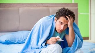 Mladý muž, který je nemocný. Sedí na posteli, přikrytý dekou a s hlavou v ruce